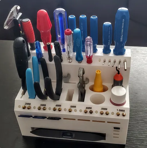 A clean, white 3D-printed workstation organizer featuring dedicated slots for pliers, hex keys, SD cards, spare nozzles, and adhesive bottles.