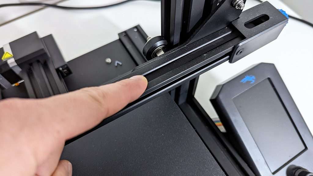 A close-up view of a finger pointing to the rubber v-roller wheels on a 3D printer's linear rail axis.