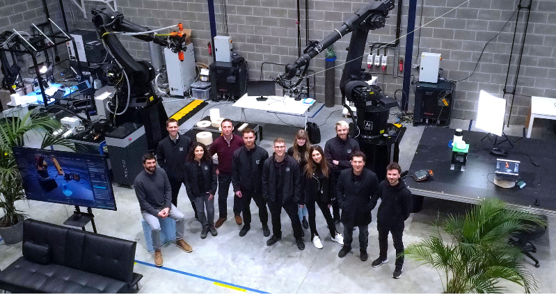A group of people posing together inside an industrial workspace featuring large robotic 3D printing arms.