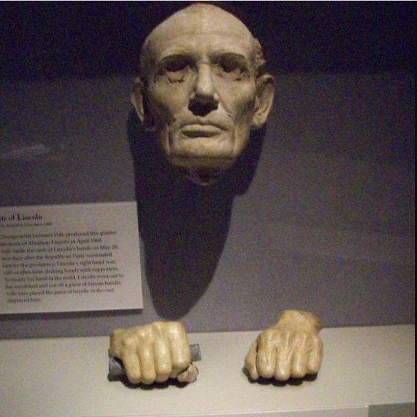 A museum display featuring a historical plaster life mask of Abraham Lincoln's face hovering above two casts of his hands.