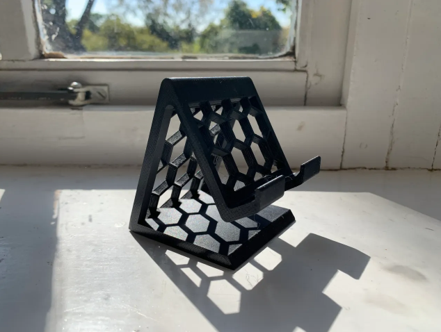 A black 3D printed universal smartphone stand featuring a hexagonal honeycomb pattern