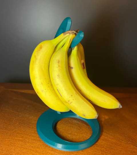 A bunch of yellow bananas hanging suspended from a minimalist, teal 3D-printed circular banana stand on a wooden surface.