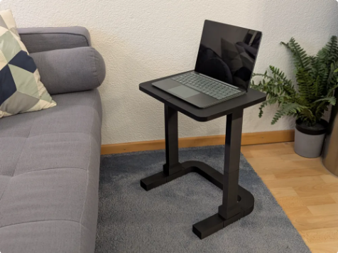A black 3D-printed modular side table supporting a laptop next to a gray sofa, designed for working from a couch or bed
