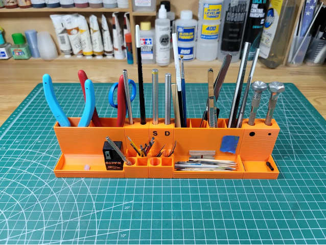 A bright orange, modular 3D-printed tray system holding X-Acto knives, brushes, drill bits, and modeling tools securely on a green self-healing cutting mat.