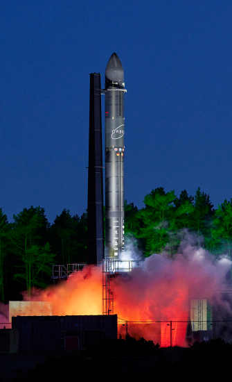Dramatic night-time launch rendering of Relativity Space's Terran R reusable rocket, lifting off with intensely bright orange exhaust flames and illuminating the launchpad structures.