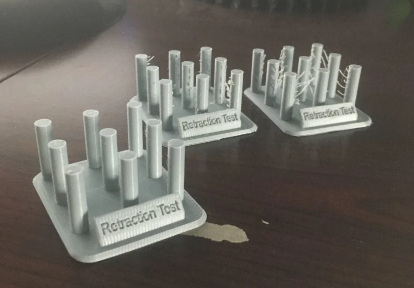 Three grey 3D-printed retraction test pillars placed side-by-side on a desk, demonstrating varying levels of stringing from a perfectly clean print on the left to one with severe web-like plastic strands on the right.