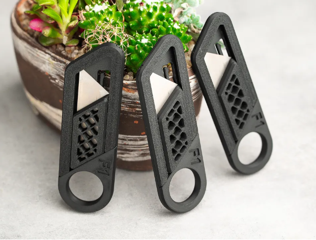 Three sleek, black 3D printed utility knife handles with exposed metal razor blades, lying on a table next to a small potted succulent.