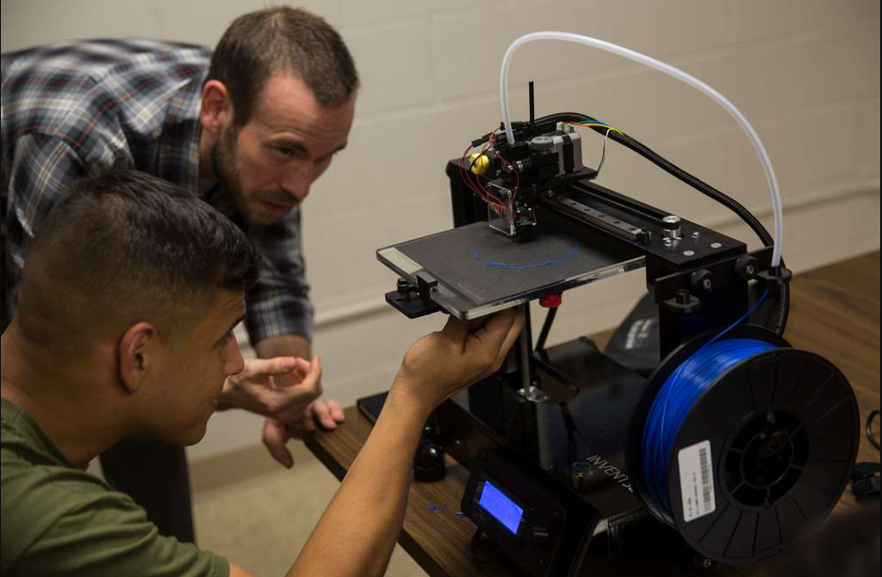 Two educators observing and adjusting an open-frame desktop 3D printer during operation.