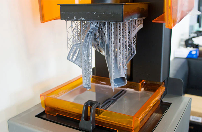 Resin 3D printer with an orange UV cover showing a complex grey 3D printed model successfully attached to the build plate