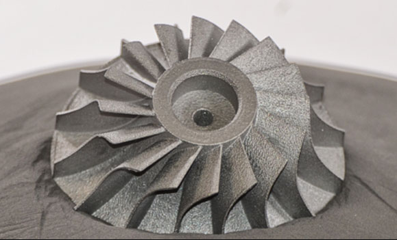 A dark grey, 3D printed metal turbine impeller part resting on a bed of recycled metal powder from 6K Additive