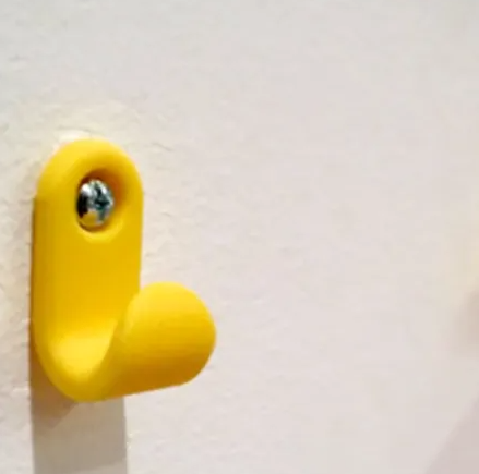 A bright yellow, minimalist 3D printed simple hook securely mounted to a white wall with a single Phillips head screw.