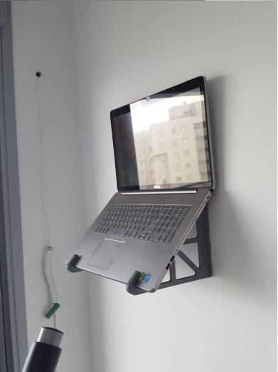 A laptop securely held by a foldable 3D-printed wall mount bracket attached to a white wall, ideal for saving desk space.