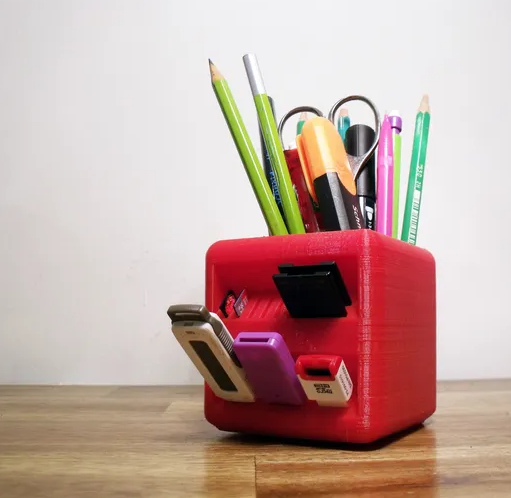 A red, 3D printed cube desk organizer known as The Little Box, storing pens, pencils, and multiple USB flash drives in specialized front slots.