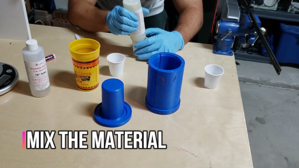 A person wearing blue nitrile gloves mixing liquid casting material in plastic cups next to a tall blue 3D-printed mold.