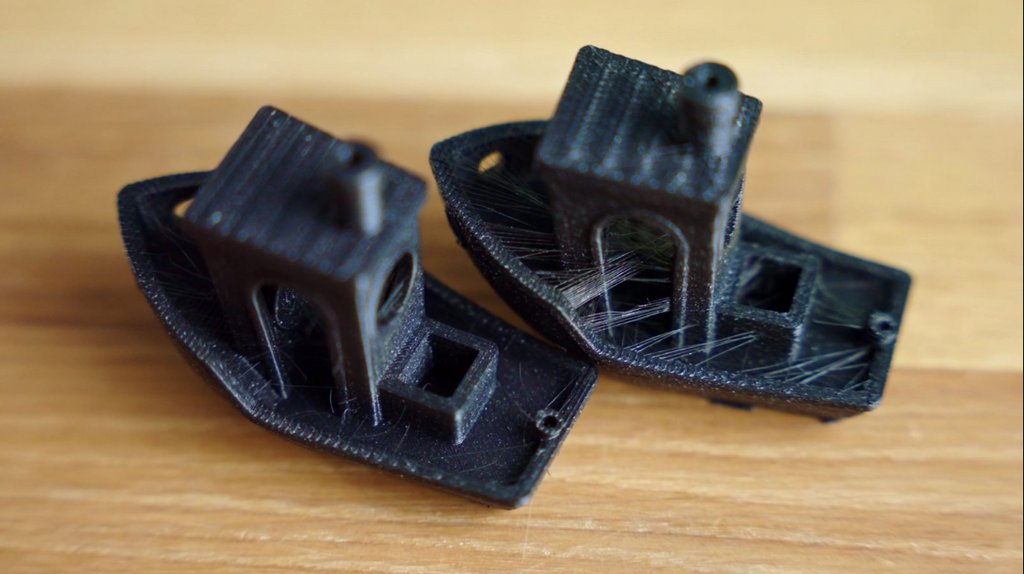 Two black 3D printed "Benchy" boat models side-by-side; the right model shows heavy stringing and surface pits caused by wet filament