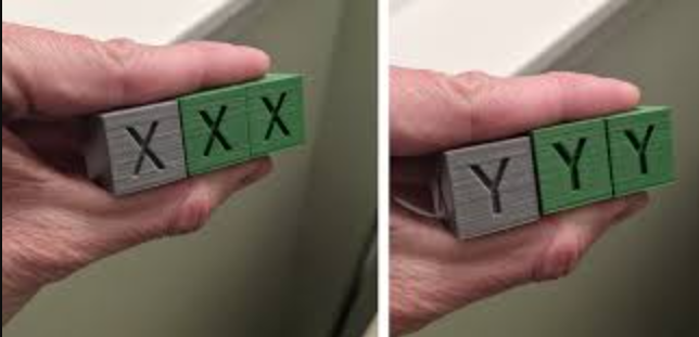 Hand holding three grey and green 3D test cubes displaying X and Y axis markings to check for surface ghosting.