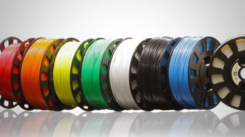 A row of colorful 3D printing filament spools, including red, orange, yellow, green, white, black, and blue, lined up on a reflective surface.