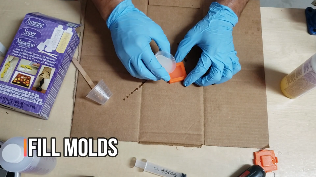 Gloved hands carefully pouring liquid casting material from a small measuring cup into an orange 3D-printed mold on a cardboard mat.