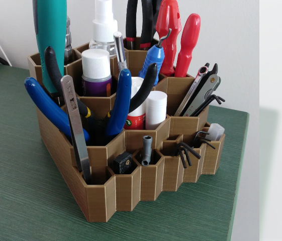 A brown, multi-tiered 3D-printed tool organizer featuring a geometric honeycomb design, filled with pliers, precision files, and craft supplies resting on a green surface.