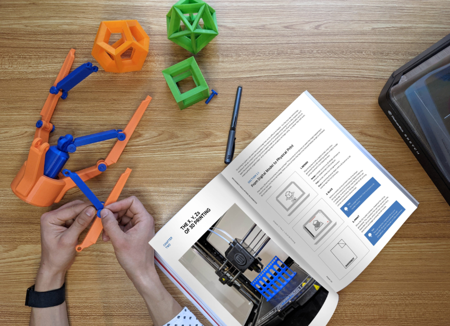 A person assembling an orange and blue 3D printed mechanical arm while following instructions from the open MakerBot Educator's Guidebook