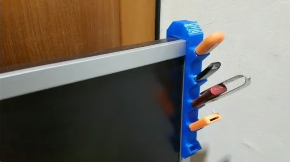 A blue 3D printed USB holder hooked vertically onto the side edge of a computer monitor, efficiently storing multiple flash drives.