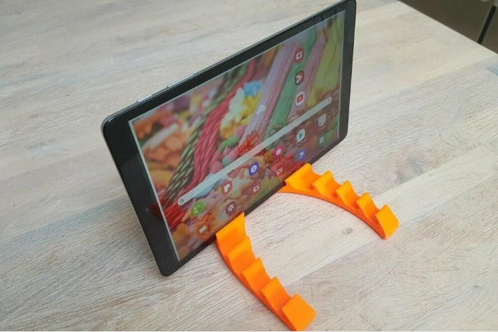 A bright orange 3D-printed tablet stand securely holding a tablet in landscape mode on a light wooden surface