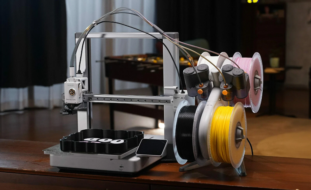 A dual-extruder 3D printer loaded with white, black, and yellow filament spools, resting on a wooden table