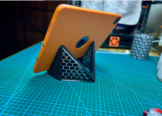 A sturdy, black 3D-printed tablet stand with a honeycomb mesh pattern supporting an orange iPad in landscape orientation on a blue grid mat