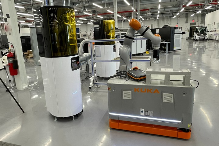 Industrial 3D printers alongside a mobile automated vehicle equipped with an articulated robotic arm.