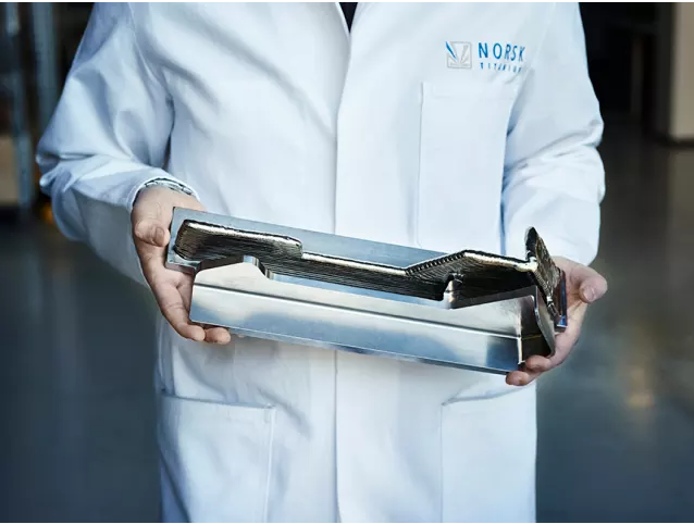 An aerospace engineer in a white lab coat holding a large, shiny, complex 3D-printed metal component for aircraft or spacecraft